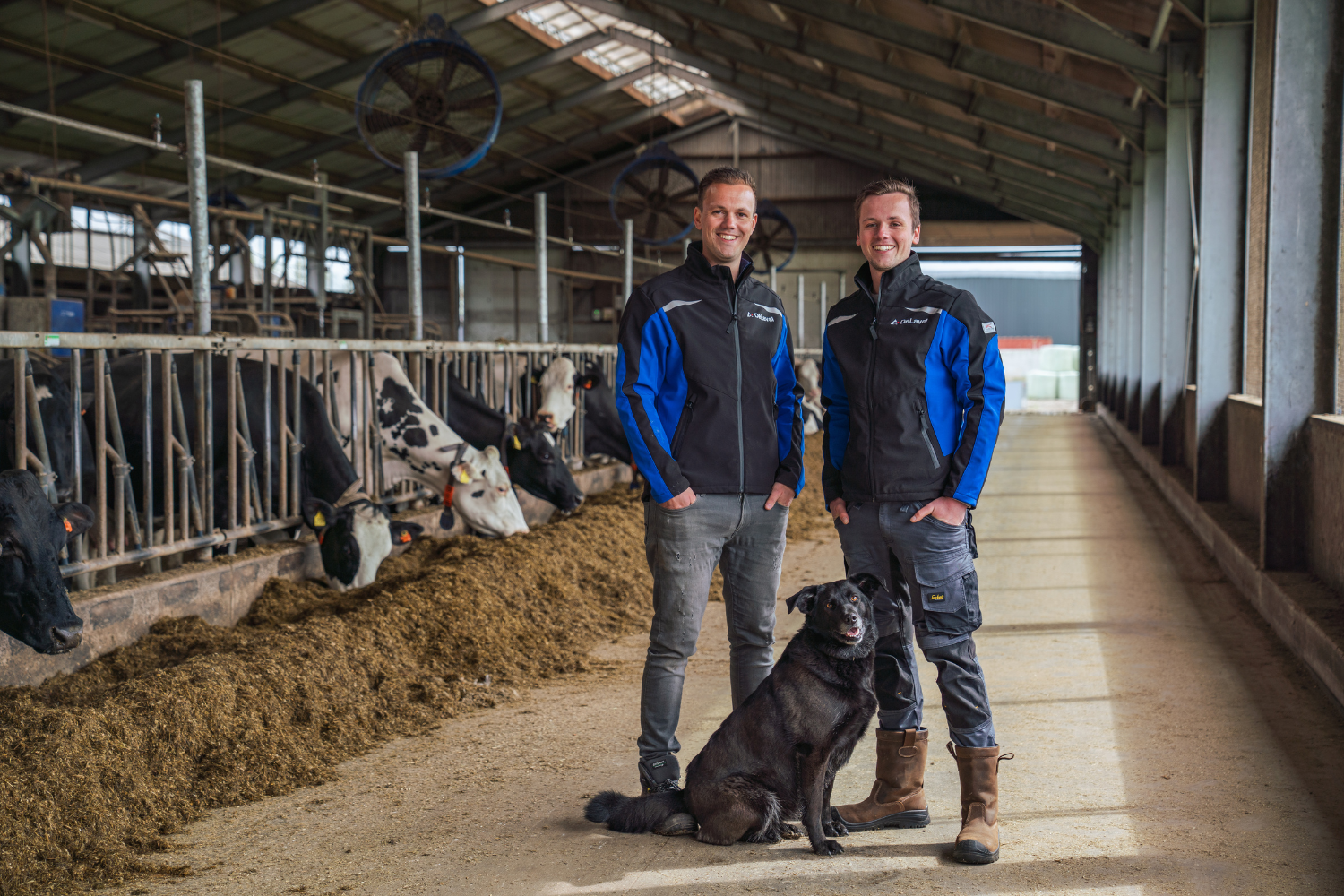 Melkveehouders Arjan en Theo van Houten met hun hond op de voergang met koeien aan het hek, beide in een jas van DeLaval